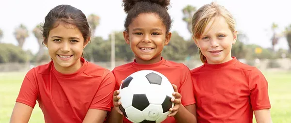 Three young girls in red jerseys smiling and holding a soccer ball, symbolizing the critical role of sports in promoting girls' mental health.