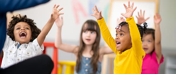 Young children sitting and raising their hands enthusiastically in a classroom setting