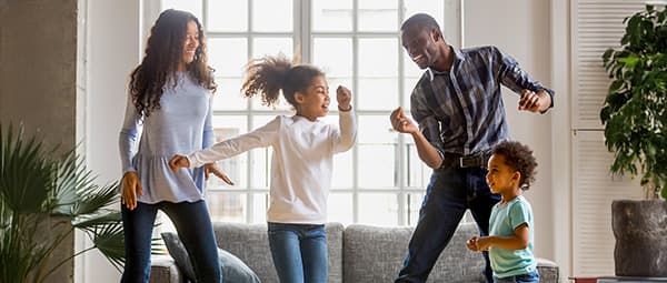 Family dancing together in a living room with large windows, looking joyful and energetic.