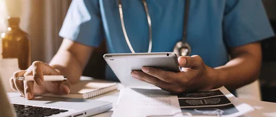 A doctor's hands holding a tablet and writing on a clipboard.