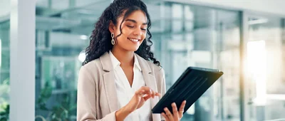 A professional woman in a light-colored blazer using a tablet in a modern glass office environment, smiling and looking down at the device.