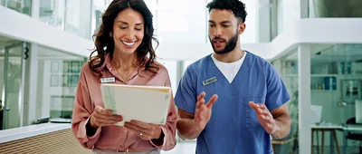 Two healthcare professionals standing in a modern medical facility, one in a pink blouse holding a tablet, the other in blue medical scrubs gesturing while talking.