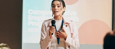 A person speaking into a microphone at a conference or presentation, wearing a white jacket and lanyard, with a blurred background showing presentation slides.
