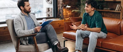 A counselor or therapist listens to a younger client in a session, illustrating a discussion or consultation in a professional yet relaxed environment.