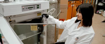 A student in a lab coat and gloves is placing a sample into a scientific incubator.