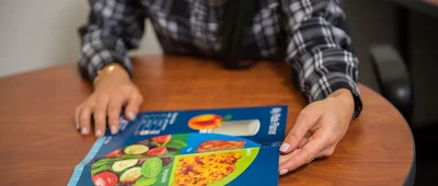 A cookbook or recipe book open on a wooden table, with a person in a plaid shirt sitting nearby.
