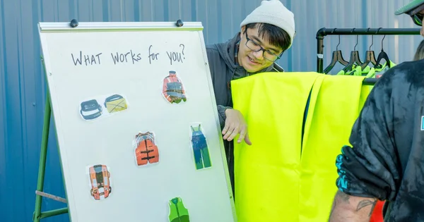 A person in a yellow safety vest standing next to a whiteboard displaying various safety equipment icons.