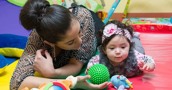 A woman interacting with a young child, both surrounded by colorful toys and cushions.