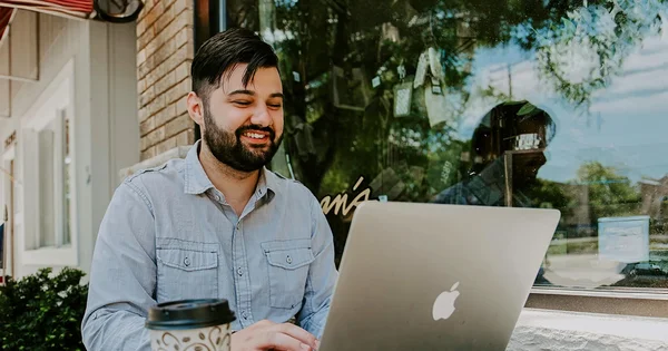 A smiling man with a beard sitting outdoors and using a laptop.