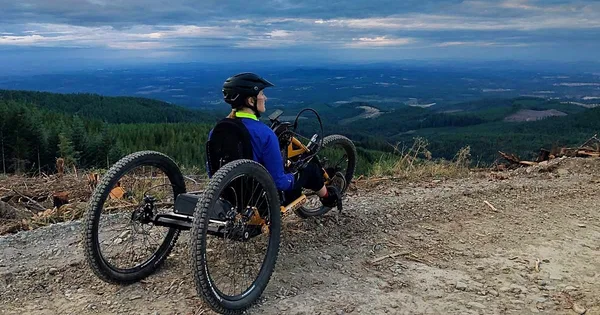 A person riding a specialized three-wheeled bicycle on a dirt trail in a mountainous, forested area.