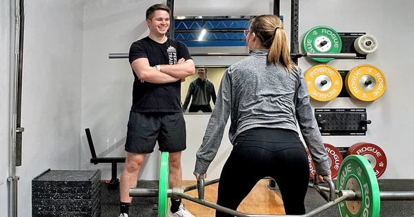 Two people in a gym setting, one standing and one lifting weights.