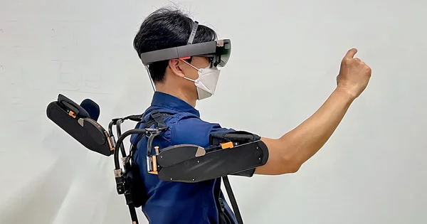 Person wearing augmented reality headset and exoskeleton arm support, viewed from the side against a white background.