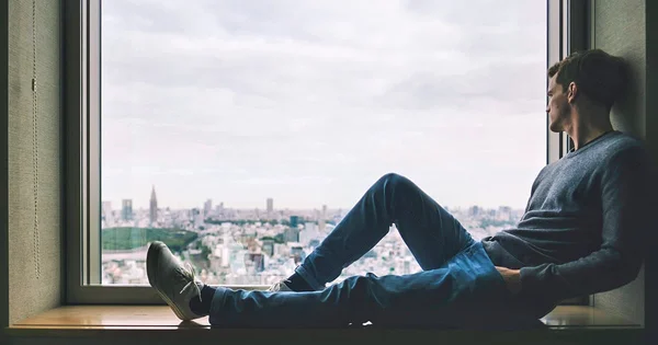 A person sitting on a windowsill, looking out over a city skyline, wearing dark clothing and shoes.