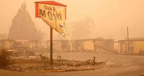 Devastated landscape with a damaged sign, burned buildings, and a destroyed car in an orange-tinted, smoke-filled environment.