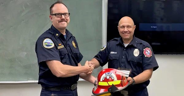 Two emergency service workers in dark blue uniforms shaking hands, with a red firefighter helmet in the foreground.