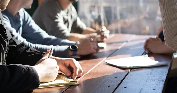 People sitting at a wooden table, taking notes during a meeting or study session.