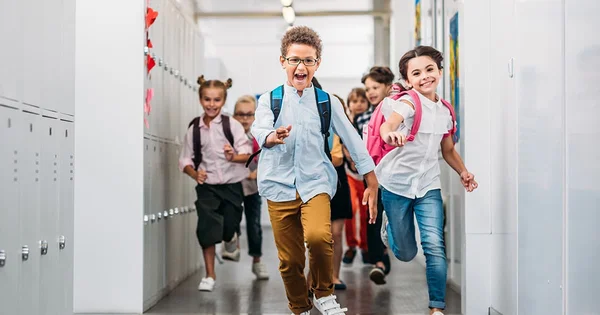 Excited school children running in a hallway with lockers, wearing backpacks and school clothes.
