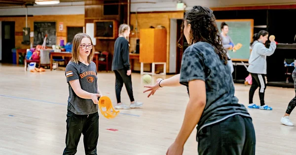 Students playing with ball in a gymnasium or activity room, with multiple people in the background.