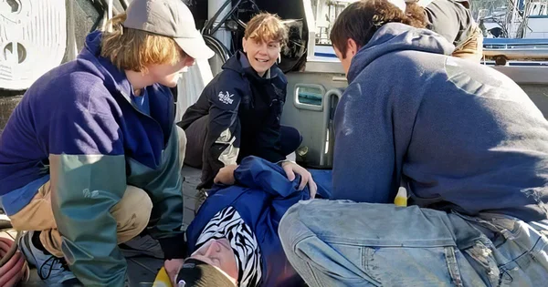 Three people in blue jackets sitting on a boat deck, one person appears to be receiving medical attention or first aid.