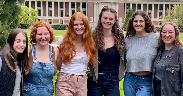 Six young women standing together on a campus lawn with a brick building in the background, smiling and posing for a group photo.