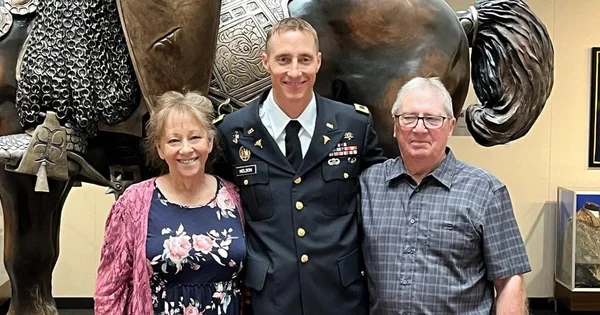 Three people posing together in front of large sculptural artwork, with a military officer in dress uniform standing between an older woman and an older man.