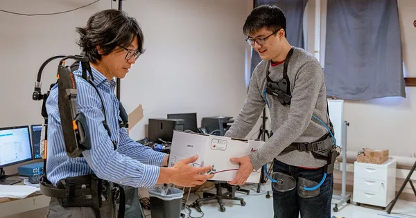 Two people in a technical workspace, wearing exoskeletons or support devices, handing a heavy box from one to another.