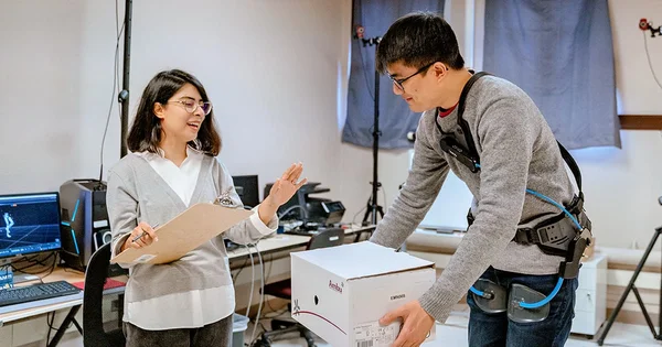 Two people in a technical workspace, with computer monitors and equipment, interacting near a white box.