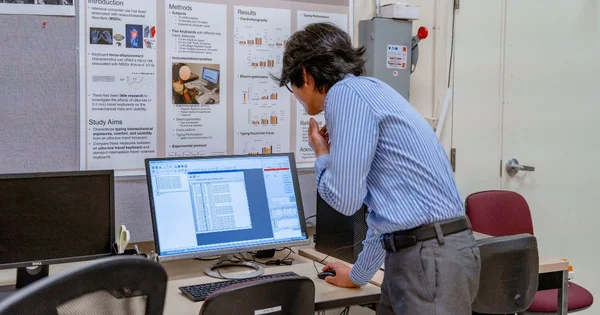 Researcher working at a computer with research poster in background, wearing a blue and white striped shirt.