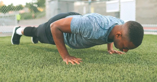 An athlete performing a push-up exercise on an artificial grass surface, wearing a blue-gray athletic shirt and black pants.