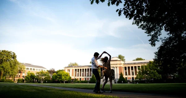 Two people dancing on a college campus pathway with historic buildings in the background, surrounded by trees and green lawn.
