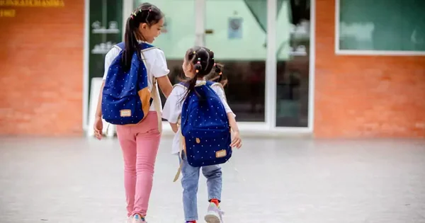 Two school-aged children with backpacks walking away from the camera near a school building.