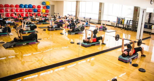 Fitness class with multiple people exercising on step platforms in a bright gym with exercise balls and weights.