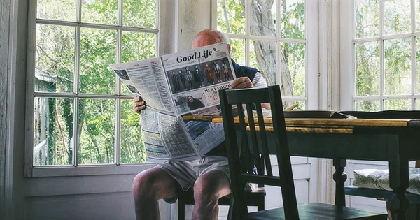 An older individual reading a newspaper by a bright, tree-lined window, sitting at a wooden table with chairs.