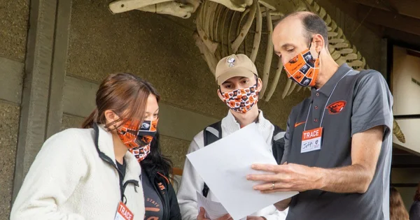 Three people wearing masks and university-branded clothing, standing near a skeletal display in an indoor setting.