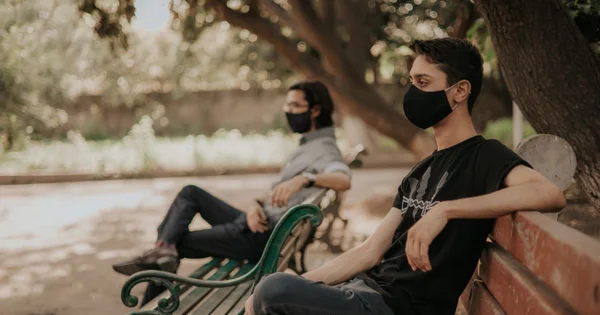 Two people wearing black face masks sitting on a park bench under trees, socially distanced from each other.