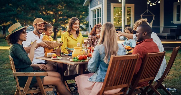 Diverse group enjoying an outdoor meal together at a wooden table with food and drinks, in a backyard setting with a house and trees in the background.