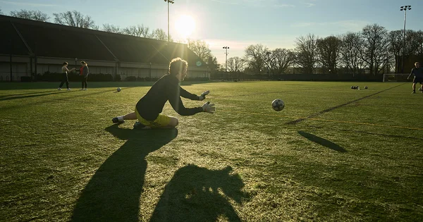 Soccer goalkeeper diving to block a ball on a sunlit field with long shadows.