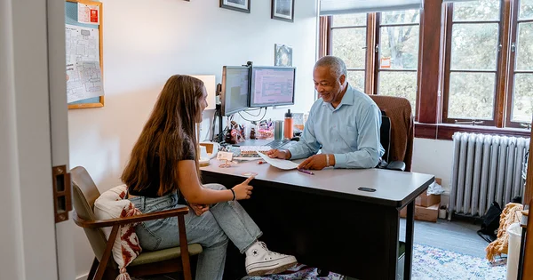 An older individual and a younger person sitting at a desk in an office with windows, engaged in conversation.