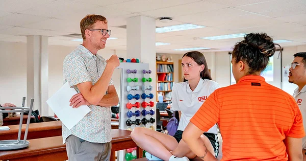 A group of people in a classroom or training room with exercise equipment and bookshelves, engaged in a discussion.