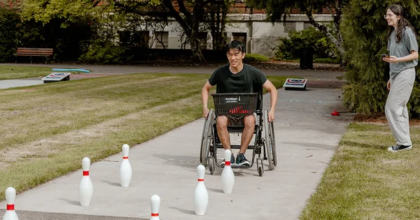 A person in a wheelchair playing bowling with white pins on a concrete path, with another person standing nearby and cornhole boards in the background.