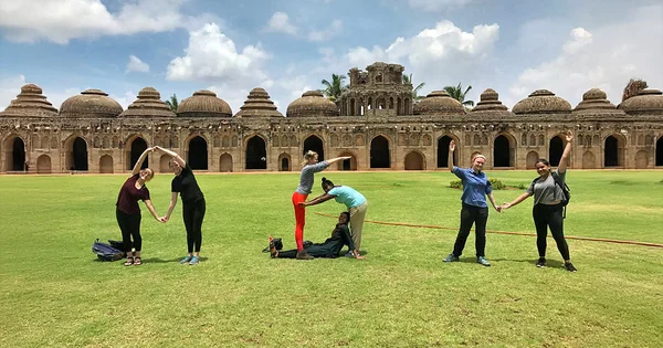 Group of people stretching and doing yoga poses on a grassy field in front of an ancient stone architectural complex with multiple domed structures.