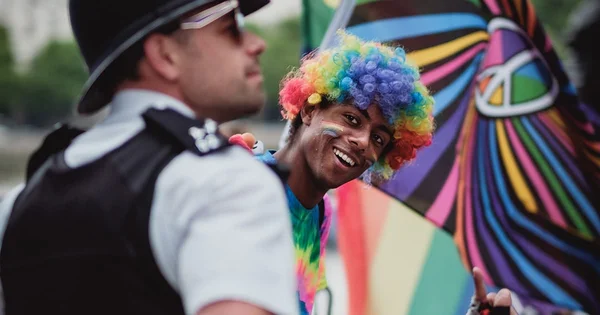 Person with rainbow-colored afro wig smiling next to a person in a formal black and white uniform, with a colorful flag in the background.