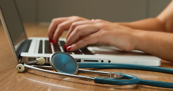 Hands typing on a laptop with a stethoscope in the foreground