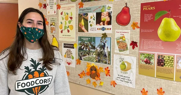 A person standing in front of a bulletin board filled with pear-themed posters and decorative autumn leaves, wearing a mask and a FoodCorps sweatshirt.