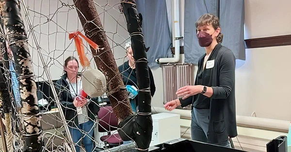 A group of people interacting with a large net structure in a classroom or workshop setting. One person is speaking while others observe, and various objects are hanging from the net.