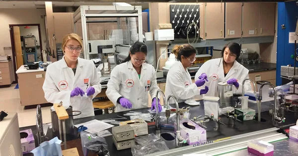 Four researchers in white lab coats and safety glasses working in a scientific laboratory with equipment and fume hoods.
