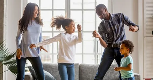 Family dancing together in a living room with large windows, looking joyful and energetic.