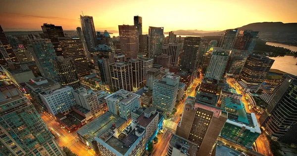 Aerial view of a dense urban cityscape at sunset with skyscrapers, illuminated streets, and mountains in the background.
