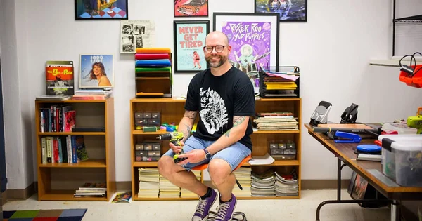 Sam Logan wearing a t-shirt, shorts, and purple sneakers, with visible arm tattoos, sits centered in a cluttered room featuring posters, vinyl records, organized supplies, and a workbench with tools.