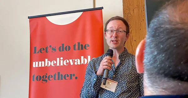 A speaker delivers a presentation or address, with an inspirational message banner behind him, suggesting a motivational or collaborative event.